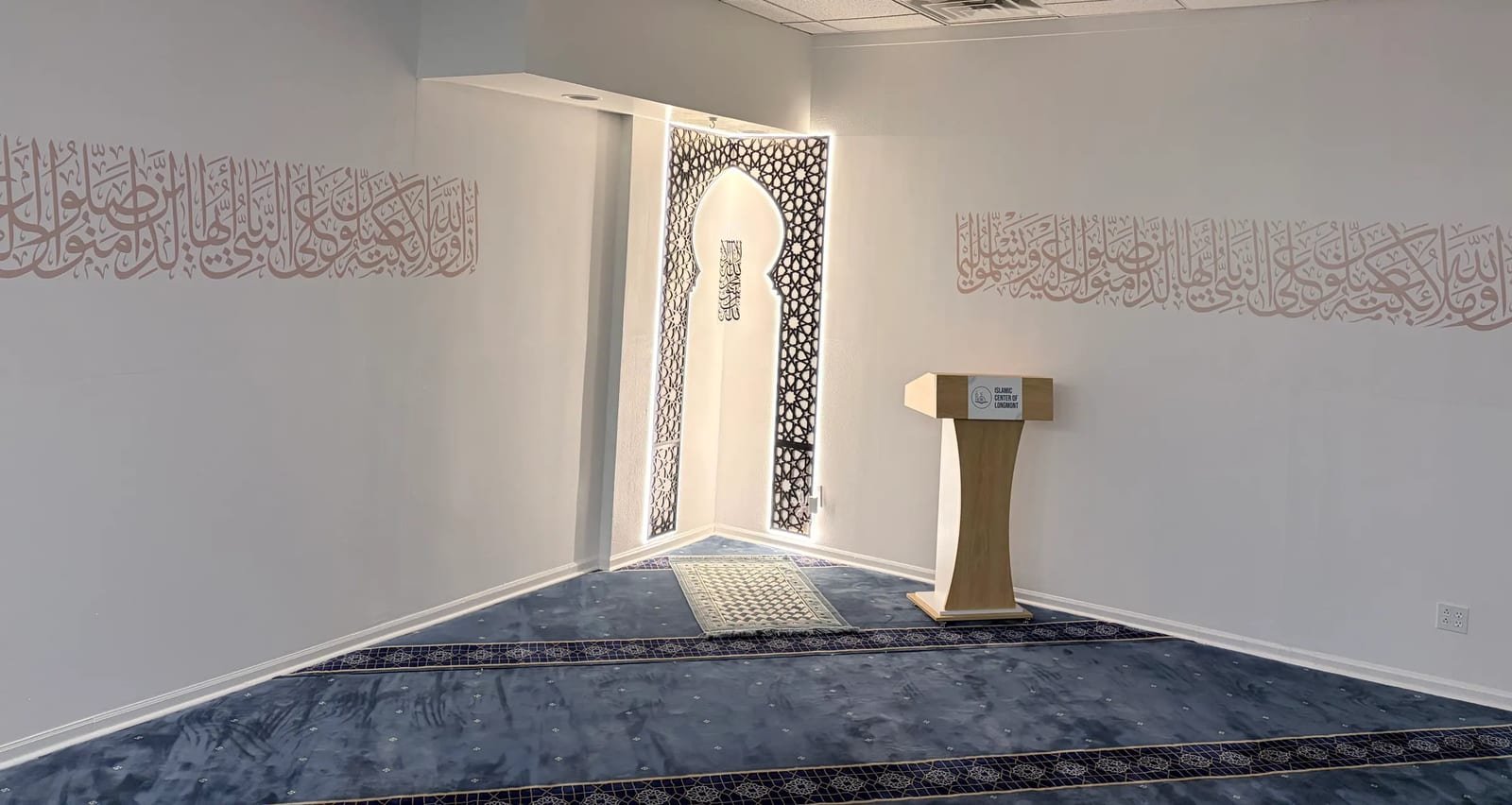 The prayer hall at Masjid Al Huda: the mihrab framed by a carved girih screen, with Qur'anic calligraphy along the walls and the minbar to one side.