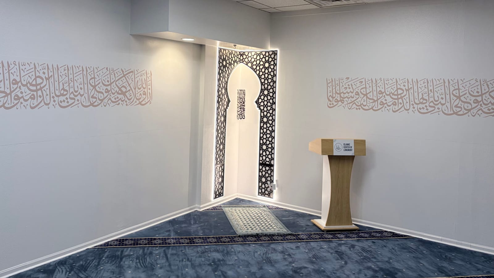 ICL's prayer hall: the mihrab in a carved girih screen, lit from within, with Qur'anic calligraphy along both walls and the wooden minbar to its right.